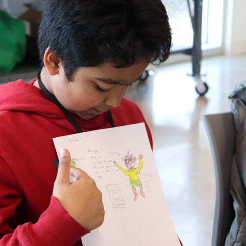 A young boy holds a piece of paper up, which has illustrations and a written story. He’s pointing to the words on the page.