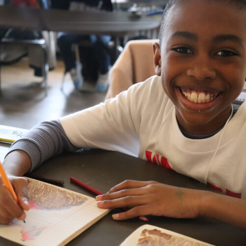 A young boy looks at the camera, smiling. He holds a pencil in his hand, colouring in a notebook.