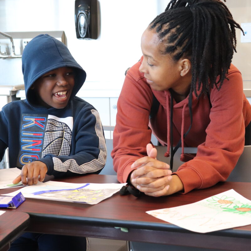 A woman leans on a desk, speaking to a young boy. The boy holds a pencil in his hand as he writes in his notebook.