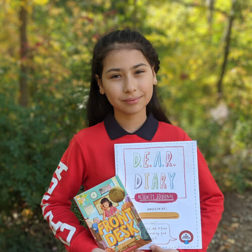 A young girl stands outside, holding a copy of the book “Front Desk” and a D.E.A.R Diary Activity Book.