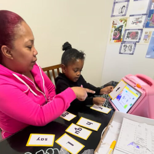 A mom sits at the table with her daughter. The mom is pointing to the cue card that her daughter is holding, which has the letter ‘f’ on it.