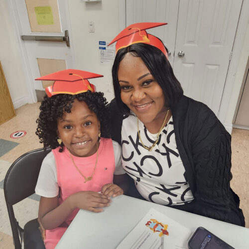 A mom and her young daughter both wear a graduate hat and smile at the camera.