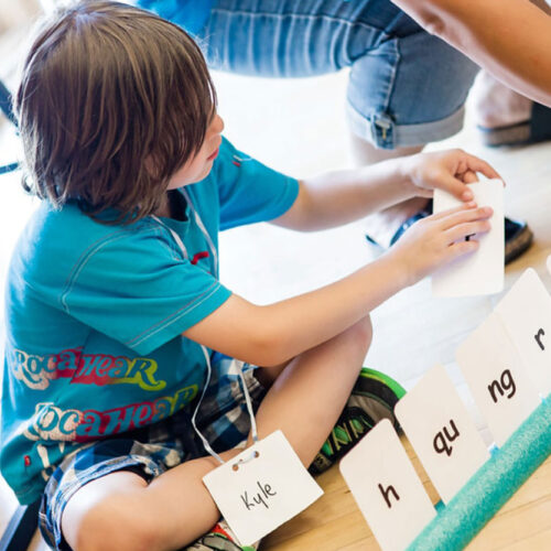 A young boy sits cross-legged on the floor. He’s holding up a cue card with a letter on it.]