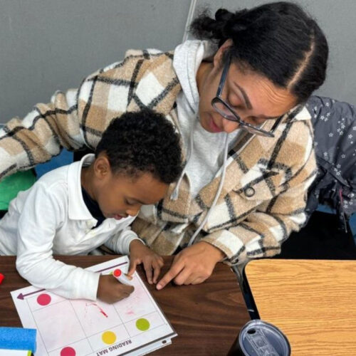 A mom and her son sit at a table. The mother is pointing to a piece of paper while her son holds a pencil.