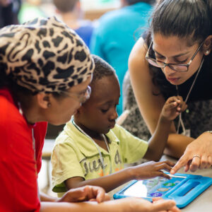 A young boy sits between two women. One woman points to the boy’s tablet, engaging in an activity with him.