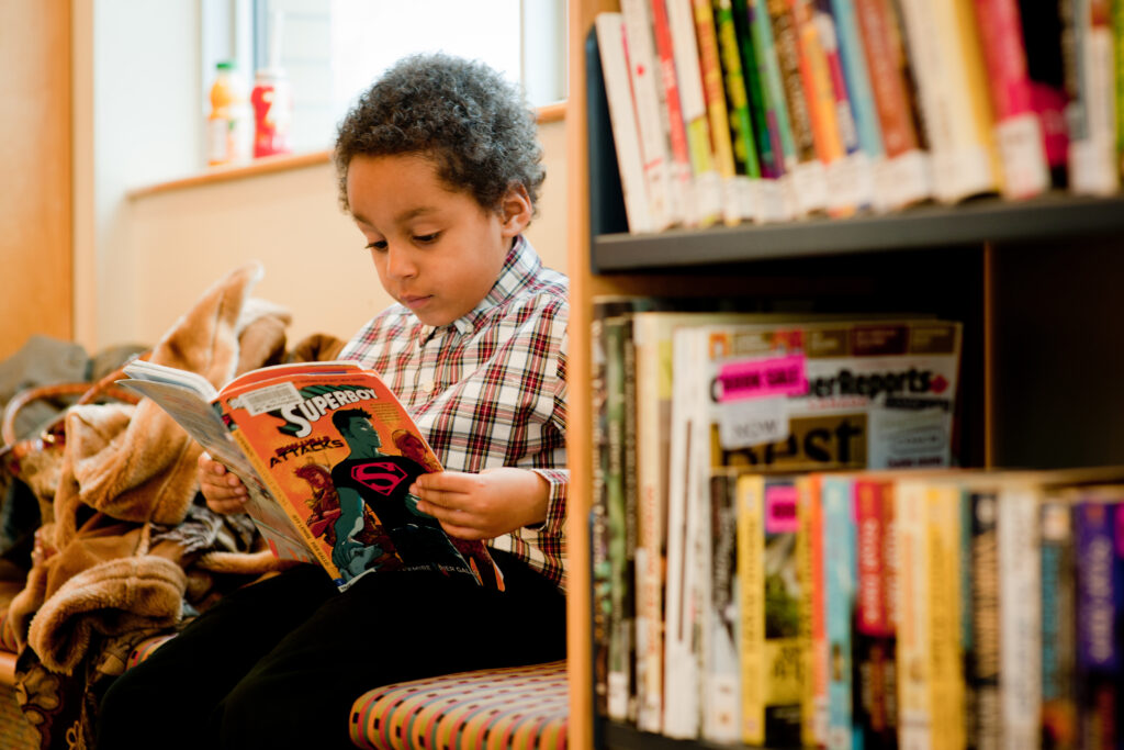A young boy sits on a bench, reading a book about superheroes on his own.